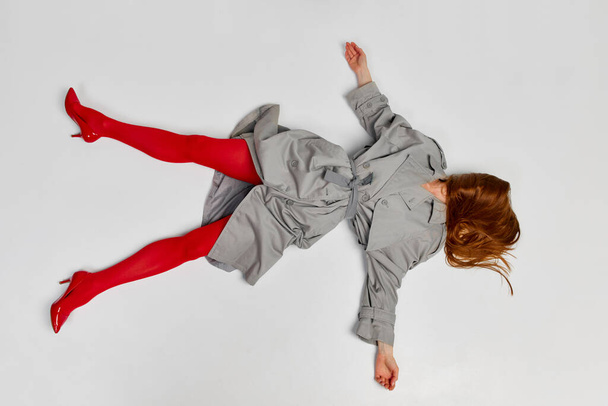 Impersonal human emotions. Stylish girl in grey coat and bright red tights moves over light background. Expressive fashion. Concept of art photography, beauty. Redhaired model in contemporary dance - Photo, Image