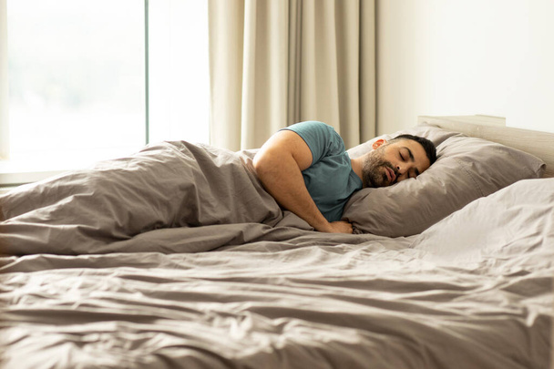 Kaukasischer Mann schläft auf der Seite im Bett, liegt unter einer Decke, schläft zu Hause, ruht friedlich im hellen Schlafzimmer drinnen, Kopierraum - Foto, Bild