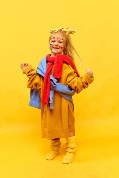 Полнометражное фото маленькой девочки в большой одежде, улыбающейся от руки к камере на жёлтом фоне. Концепция моды, имитация жизни матерей, реклама, товары для детей, детство - Фото, изображение