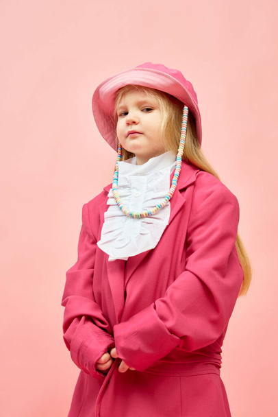 Poważna dziewczynka. Zdjęcie małej, stylowej dziewczyny stojącej i noszącej duże różowe, przerośnięte ubrania, aby naśladować matkę na różowym tle. Koncepcja mody, reklamy, dziecięcej modelki, akcesoriów - Zdjęcie, obraz