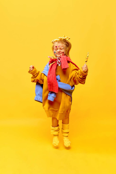 Radosne, szczęśliwe. Pełnowymiarowe zdjęcie dziewczynki, skaczącej lizakiem i uśmiechającej się z zamkniętymi oczami na żółtym tle. Pojęcie mody, reklamy, produktów dla dzieci, dzieciństwa, dziecięcej modelki - Zdjęcie, obraz