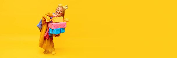 Onnellisuutta, iloa. Pikkutytöllä on paljon lahjoja, laatikoita, joissa on iloinen naama keltaisella taustalla. Käsite juhla, mainos, lasten tuotteita, lapsuus, lapsi malli. Banneri kopiotilaa - Valokuva, kuva