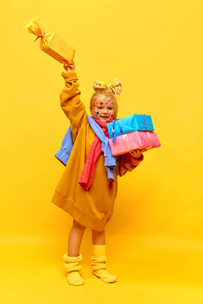 Unpacking, unboxing. Full-length photo of little girl holding gifts, showing box with happy face over yellow background. Concept of celebration, ad, childrens products, childhood, family - Photo, Image