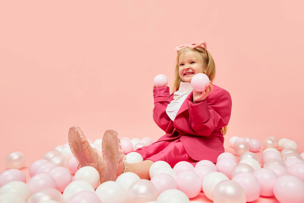 Fun, games. Photo of little girl with blond hair wearing pink clothes and sunglasses, playing with balls with hands over pink background. Concept of emotions, childhood, childrens games, ad - Fotoğraf, Görsel