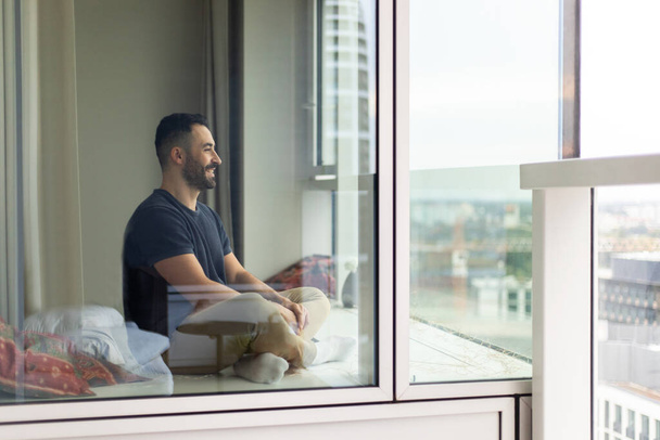 Взволнованный мужчина, сидящий на полу у окна, проводящий время в спальной зоне, наслаждающийся хорошим днем и свободным временем дома, свободным местом для копирования - Фото, изображение