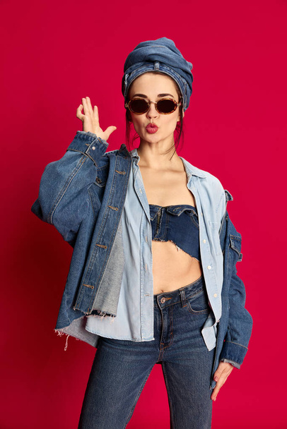 Portrait of young beautiful woman posing in stylish jeans clothes and trendy sunglasses against red studio background. Concept of extraordinary style, beauty, fashion, youth, emotions. Ad - Foto, afbeelding