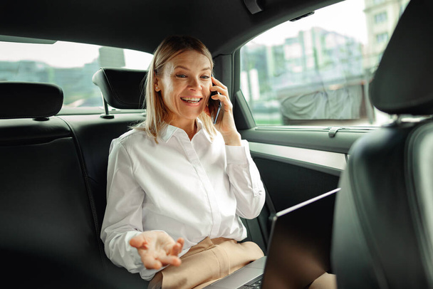 Liikenainen työskentelee kannettavan tietokoneen kanssa ja puhuu puhelimessa matkalla toimistoon autolla. Laadukas kuva - Valokuva, kuva