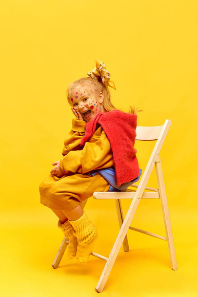 Verrast, gelukkige emoties, geluk, plezier. Foto van een lief klein meisje met strik, zittend op een stoel en lachend over een gele achtergrond. Concept van emoties, kindertijd, kindermodel, reclame - Foto, afbeelding