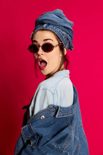 Shocked and excited face. Young beautiful woman posing in trendy stylish jeans clothes withsunglasses against red studio background. Extraordinary style, beauty, fashion, youth, emotions concept. Ad - Photo, image