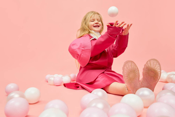 Geluk, plezier. Foto van een klein kind met blond haar in roze kleren spelend, bal gooiend en lachend over een roze achtergrond. Concept van emoties, kindertijd, kinderspelletjes, reclame - Foto, afbeelding