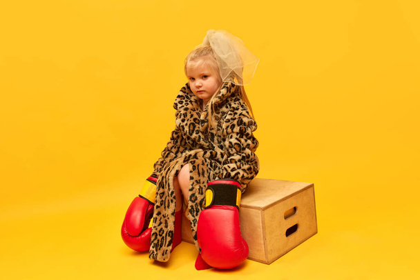 Expresión triste y perturbada. Pequeña chica rubia molesta con guantes de boxeo y elegante abrigo de leopardo sentado en cajas y mirando hacia otro lado sobre el fondo amarillo. Concepto de emociones humanas, moda, anuncio - Foto, imagen