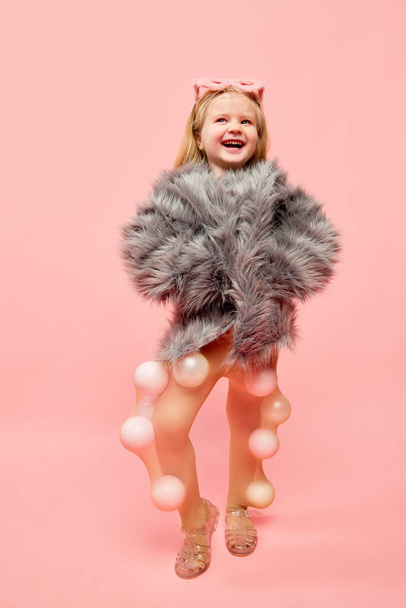 Joyful. Photo of little happy girl wearing sunglasses and fur coat, keeping hands in pocket with happy face looking away over pink background. Concept of emotions, fashion, ad, child model, childhood - Photo, Image