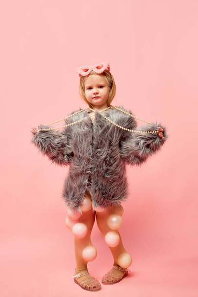 Full-length photo of little stylish blond girl wearing fur coat with sunglasses, touching necklaces, standing and looking at camera over pink background. Concept of emotions, fashion, ad, child model - Photo, Image
