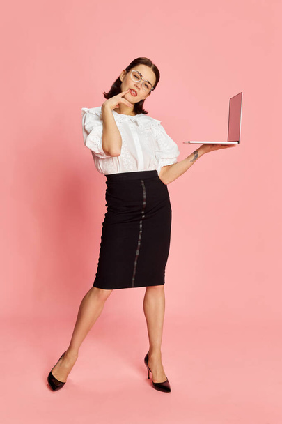 Full-length portrait of young beautiful business woman in formal wear posing with laptop against pink studio background. Concept of business, education, occupation, emotions, fashion. Ad - Zdjęcie, obraz