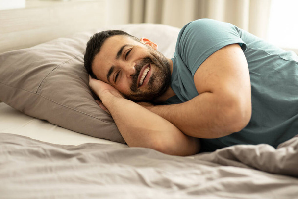 Yatakta uzanmış, kameraya bakıp gülümseyen, sabah yatak odasında dinlenen mutlu bir adamın portresi. Yüksek kalite fotoğraf - Fotoğraf, Görsel