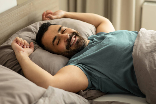 Šťastný uvolněný muž ležící v pohodlné posteli, užívající si dobré ráno v ložnici a protínající se po probuzení. Kvalitní fotografie - Fotografie, Obrázek