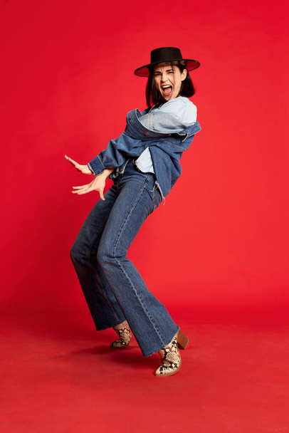 Extravagant cowboy dance. Portrait of beautiful young woman in jeans clothes and black hat posing against red studio background. Concept of style, beauty, fashion, youth, emotions. Ad - Foto, afbeelding