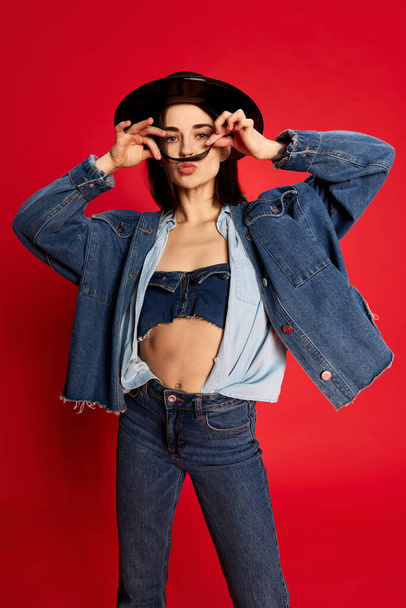 Happy, relaxed and delightful. Portrait of beautiful young woman in jeans clothes and black hat posing against red studio background. Concept of style, beauty, fashion, youth, emotions. Ad - Fotoğraf, Görsel