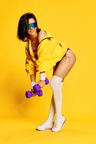 Full-length image of beautiful young woman in stylish sportswear and sunglasses posing with dumbbells against bright yellow studio background. Concept of sport, healthy lifestyle, fashion, beauty. Ad - Fotoğraf, Görsel