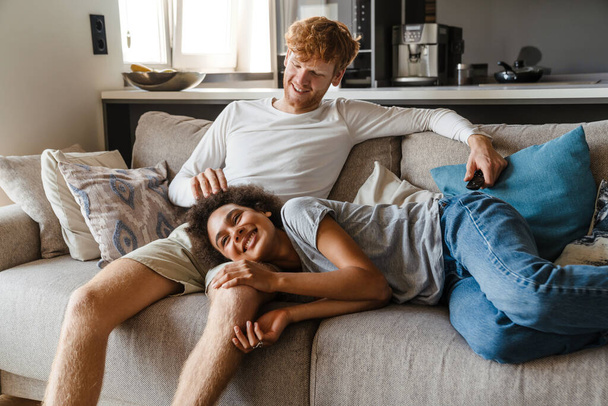 Young handsome smiling redhead guy caressing his beautiful african girlfriend liying on his knee, while they watching tv on couch at home together - Photo, Image