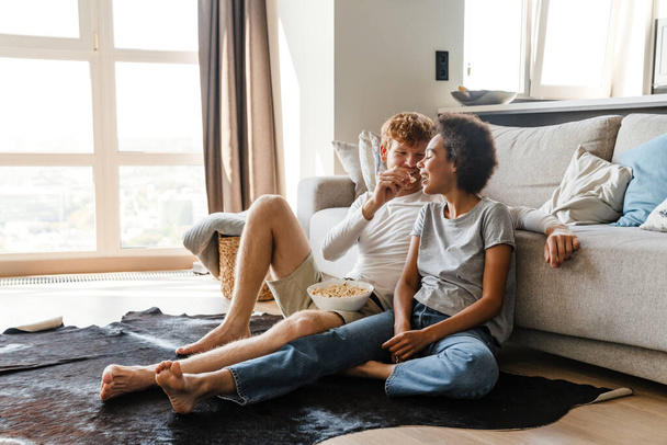 Young handsome redhead guy feeding his african girlfriend popcorn, while they sitting on the floor leaning on couch in cozy sunny living room at home - Fotó, kép