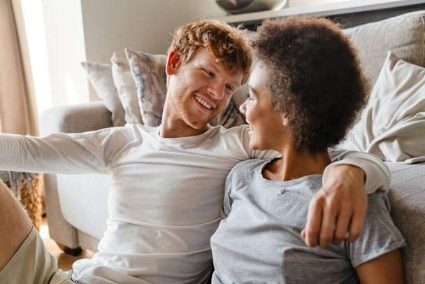 Young beautiful happy smiling interracial couple looking at each other, while sitting on the floor leaning on couch. Handsome redhead guy and beautiful african curly girl - Valokuva, kuva