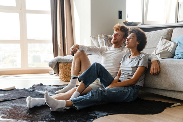 Young beautiful happy smiling interracial couple watching tv , while sitting on the floor leaning on couch at home together. Handsome redhead man and beautiful smiling african woman - Foto, Imagen