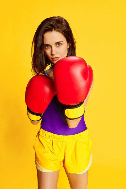 Fitness as hobby. Beautiful young brunette woman in stylish sportswear posing with red boxing gloves against bright yellow studio background. Concept of sport, healthy lifestyle, fashion, beauty. Ad - Photo, Image