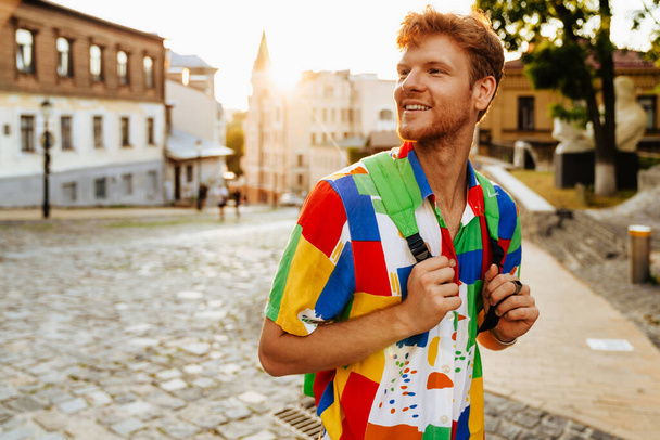 Young handsome smiling redhead man with stubble in colorful shirt holding backpack and looking aside, while standing on the street - Fotografie, Obrázek