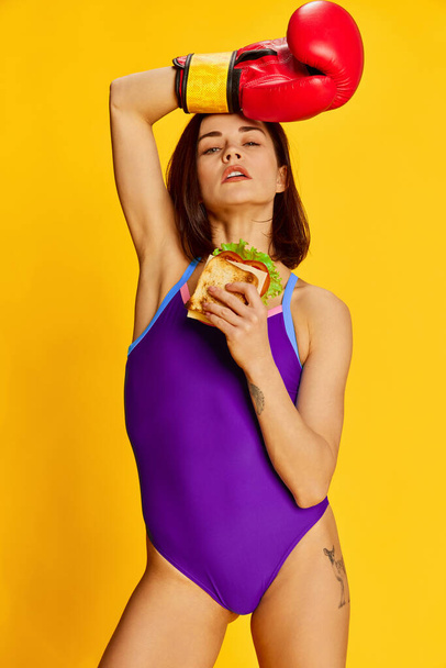 Sandwich or diet. Beautiful young brunette woman in stylish sportswear posing in red boxing gloves against bright yellow studio background. Concept of sport, healthy lifestyle, emotions, fashion. Ad - Valokuva, kuva