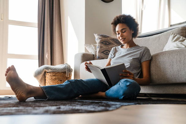 Young curly woman wearing jeans reading book while sitting on floor at home - Photo, Image