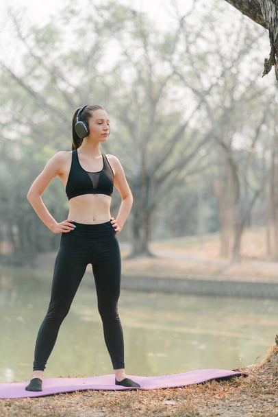 In een achtergrond van het Herfst Stadspark glimlacht een prachtige sportvrouw in sportkleding terwijl ze haar lichaam uitrekt en flexibiliteitsoefeningen doet als opwarming voor een hardlooptraining. - Foto, afbeelding