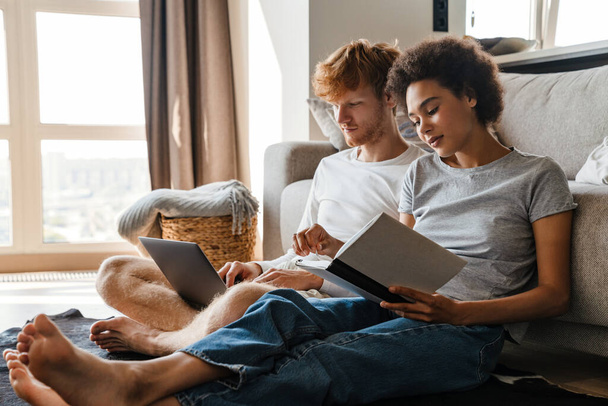 Young multinational couple using laptop and reading book while resting at home - Photo, image