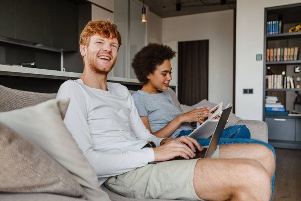 Junges multinationales Paar benutzt Laptop und Buch, während es sich zu Hause auf dem Sofa ausruht - Foto, Bild