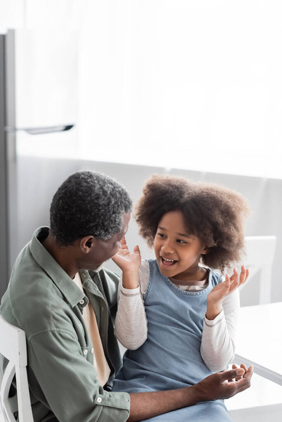 excited african american girl with curly hair gesturing while talking with cheerful grandfather at home  - Fotoğraf, Görsel