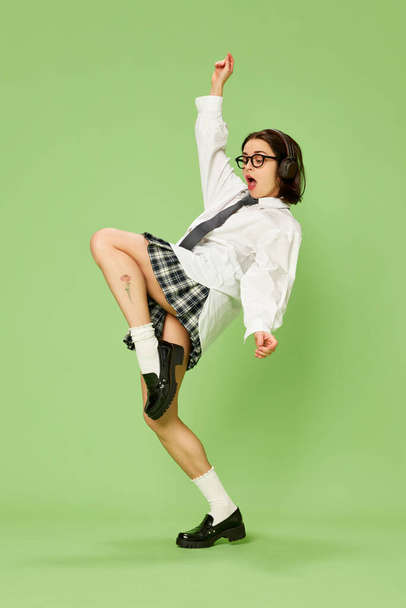 Beautiful woman in checkered skirt and white shirt posing, emotionally listening to music in headphones against green background. Concept of youth, fashion, emotions, lifestyle, facial expression. Ad - Photo, Image