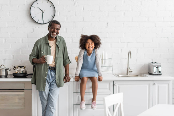 hombre afroamericano de mediana edad sosteniendo taza con café cerca de la nieta feliz sentado en encimera de la cocina  - Foto, Imagen