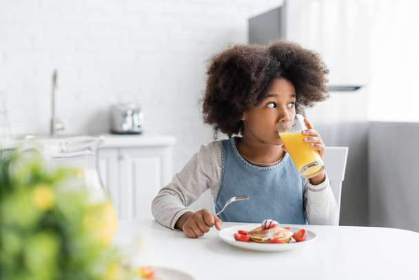 rizado afroamericano chica bebiendo jugo de naranja y sosteniendo tenedor cerca de panqueques mientras desayuna  - Foto, imagen