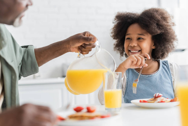 happy african american girl looking at grandfather pouring orange juice during breakfast  - Fotografie, Obrázek