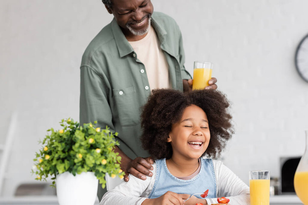 szczęśliwy Afrykański dziadek trzyma szklankę soku pomarańczowego i stoi za radosnym dzieckiem podczas śniadania  - Zdjęcie, obraz