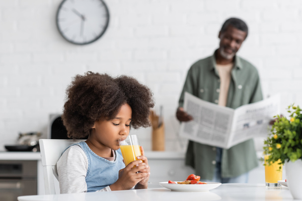 göndör afro-amerikai lány narancslevet iszik, míg nagyapa újságot olvas a konyhában - Fotó, kép