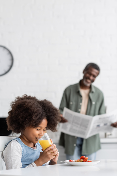 祖父の近くでオレンジジュースを飲む幸せなアフリカ系アメリカ人の子供が背景のぼやけた新聞を読んで  - 写真・画像