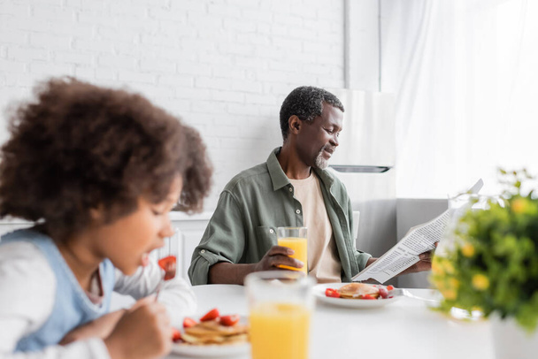 happy african american grandparent reading newspaper while granddaughter having breakfast in kitchen  - Фото, изображение