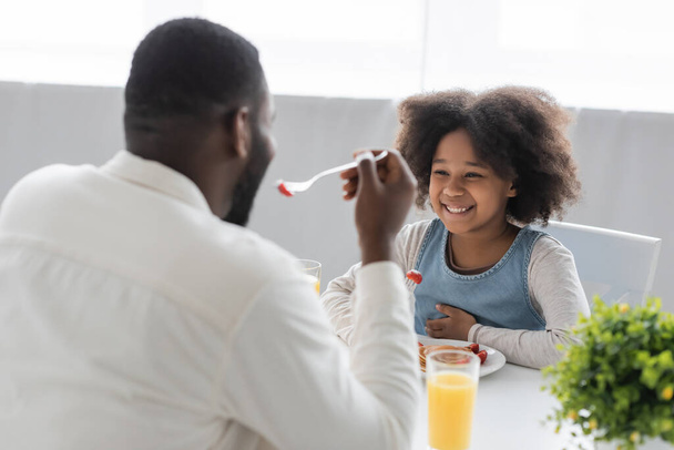 feliz afroamericana chica desayunando y mirando a padre comiendo fresa en borrosa primer plano  - Foto, imagen