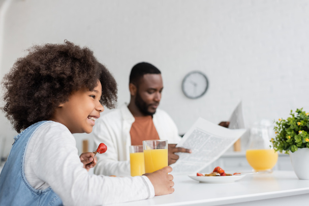 feliz afroamericana chica desayunando mientras padre leyendo periódico sobre fondo borroso  - Foto, imagen
