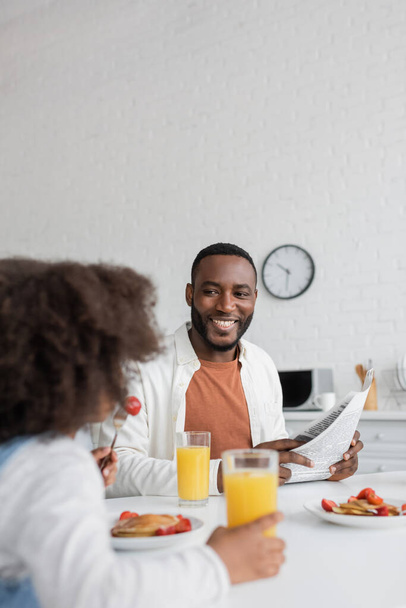 heureux homme afro-américain tenant journal et regardant fille frisée prendre le petit déjeuner au premier plan flou  - Photo, image
