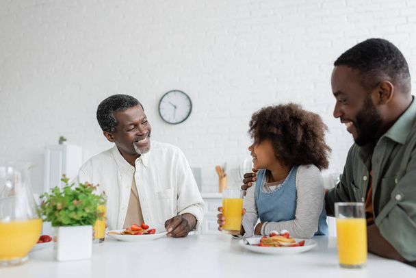 happy middle aged man looking at african american granddaughter near son during family breakfast  - Zdjęcie, obraz