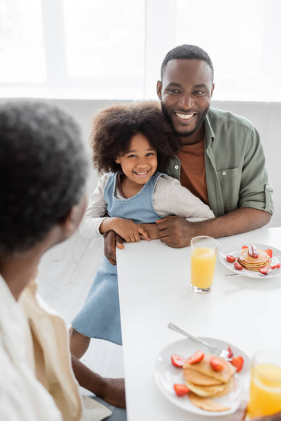 happy african american man hugging curly daughter and looking at father during family breakfast  - Foto, afbeelding