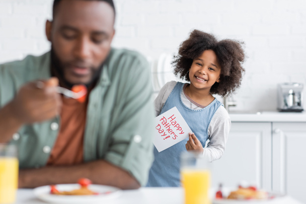alegre afroamericana chica sosteniendo tarjeta de felicitación con feliz día padres letras mientras que de pie detrás de papá en borrosa primer plano  - Foto, imagen