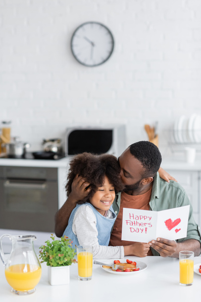 africano americano hombre celebración de tarjeta de felicitación con feliz padres día letras mientras besos cabeza de rizado hija  - Foto, imagen